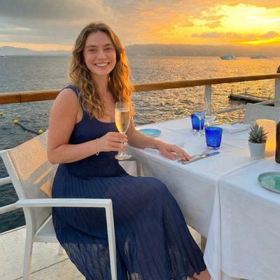 Woman in a blue dress holding a glass of wine at a sunset table by the water.